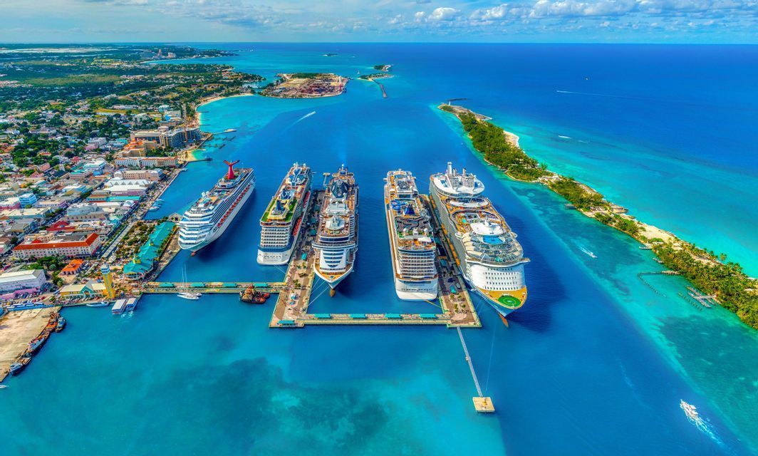 Una vista aerea di cinque grandi navi da crociera attraccate in fila a un molo in un porto tropicale con acqua turchese, vicino a una città.