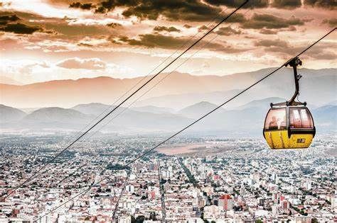 Una funivia gialla si muove su cavi ad alta quota sopra una città, con una catena montuosa visibile in lontananza sotto un cielo drammatico.