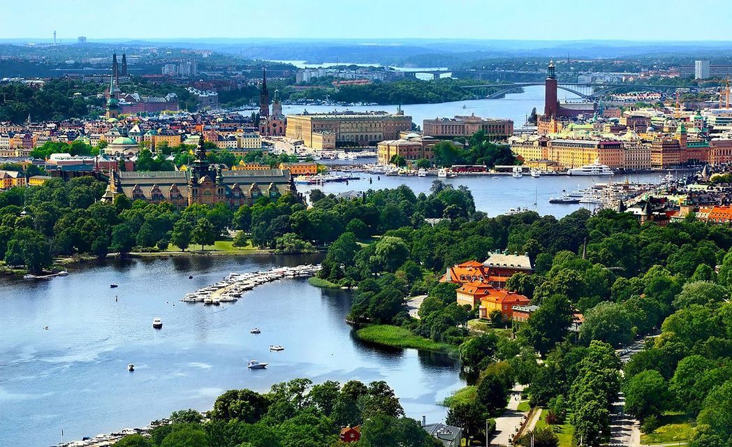 Una vista aerea di una città tentacolare lungo un corso d'acqua, con edifici storici, alberi verdi e numerose barche in una giornata limpida.