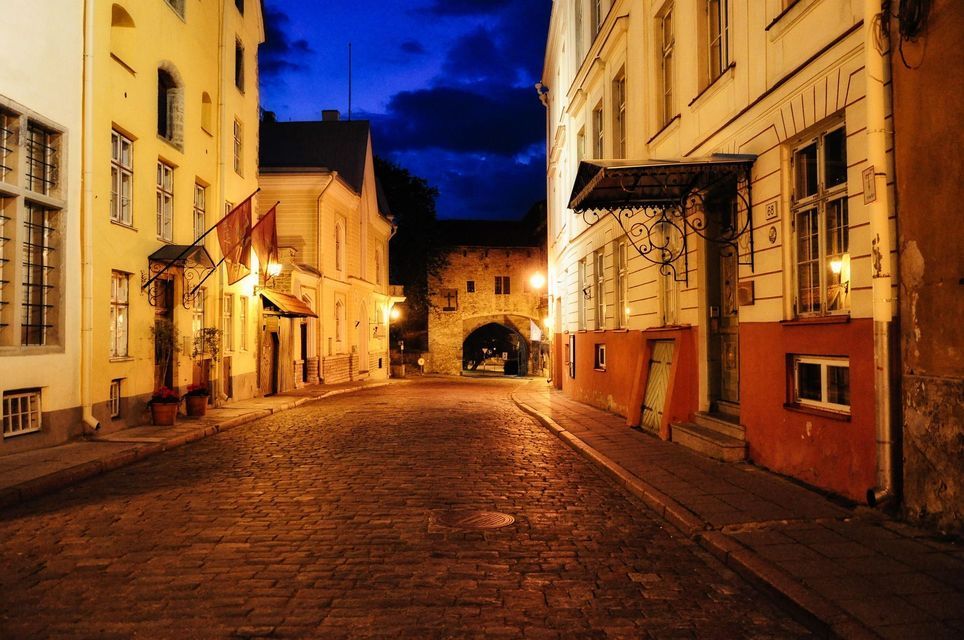 Una strada acciottolata vuota illuminata di notte, affiancata da vecchi edifici e che conduce a un arco in pietra sotto un cielo blu scuro.