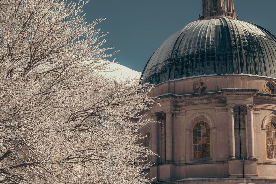 Un edificio classico con cupola si erge accanto a rami innevati, con una montagna imbiancata sullo sfondo.
