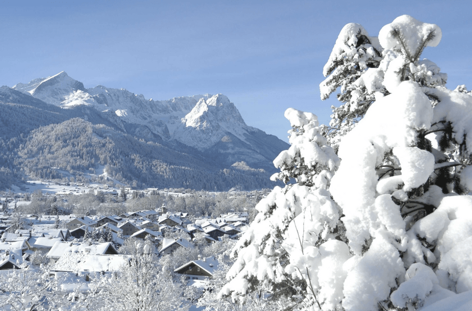 Ein schneebedecktes Dorf liegt in einem Bergtal unter strahlend blauem Himmel, der Blick geht vorbei an stark schneebeladenen Bäumen.