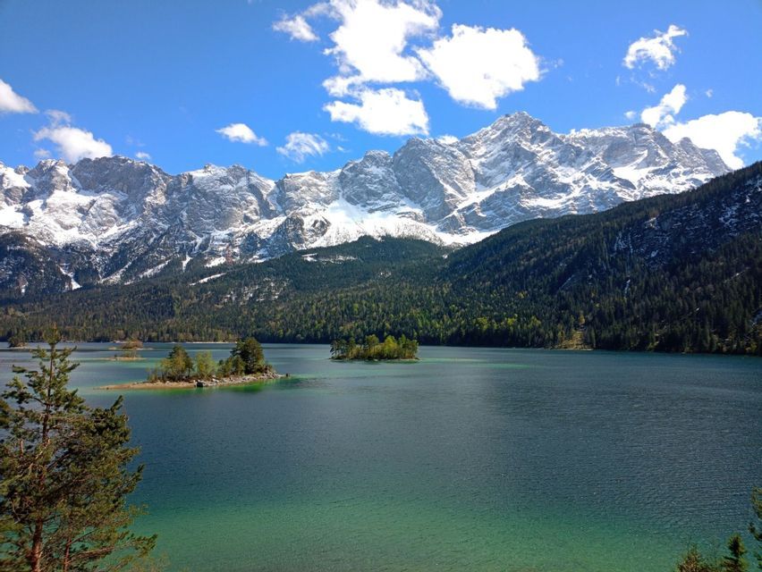 Ein türkiser See mit kleinen, bewaldeten Inseln liegt am Fuße einer schneebedeckten Bergkette unter blauem Himmel mit Wolken.