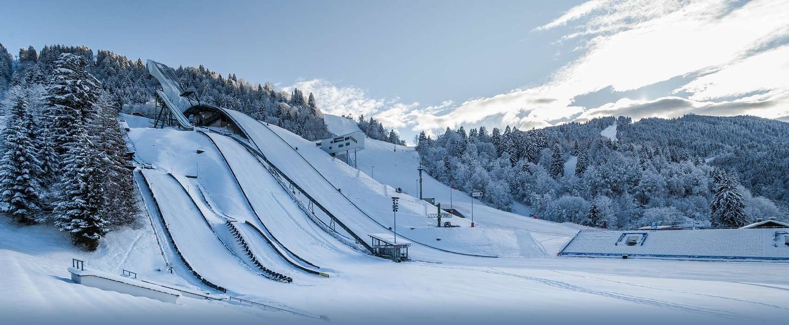 Eine große Skisprunganlage mit mehreren Schanzen an einem schneebedeckten Berghang, umgeben von Pinien.