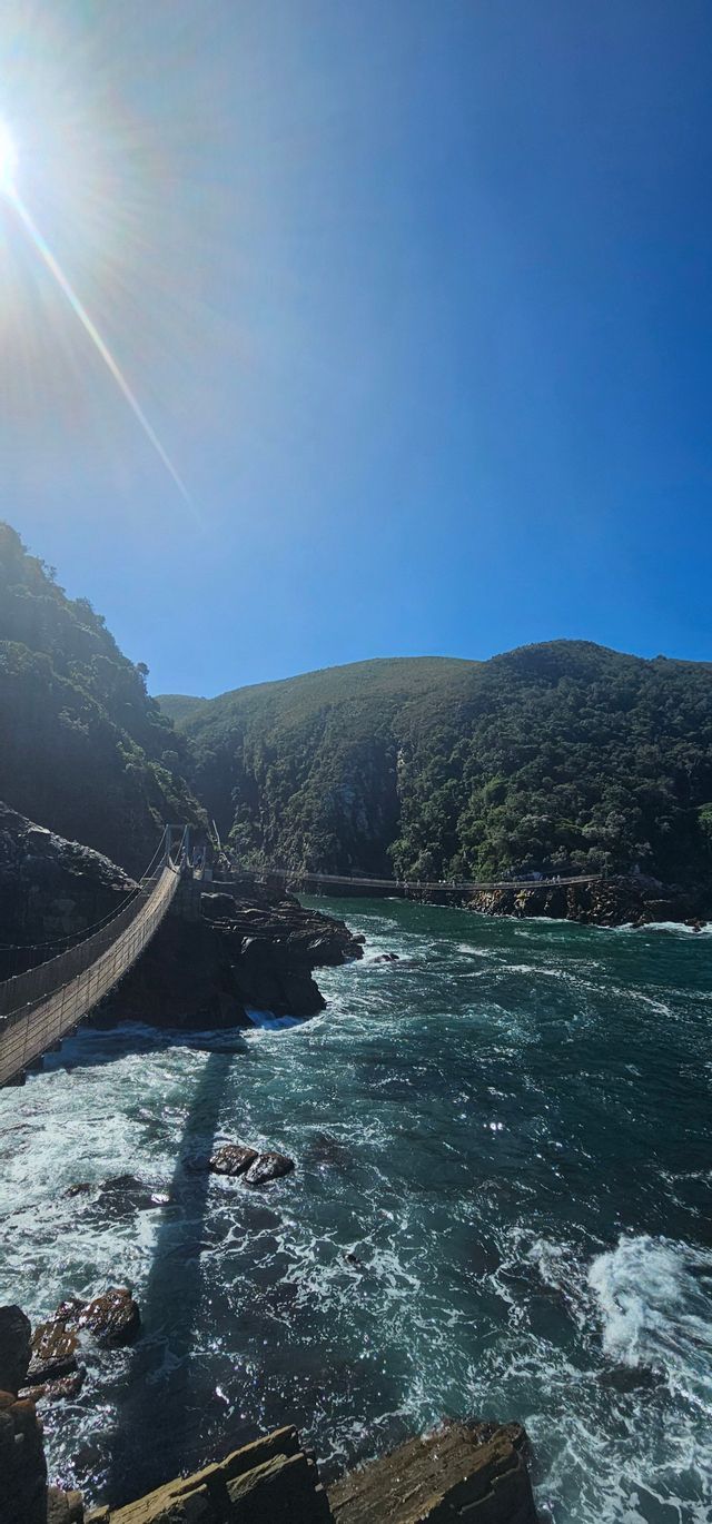 Eine Hängebrücke spannt sich an einem hellen, sonnigen Tag über tosende Küstengewässer zu dichten, grünen Hügeln hin.