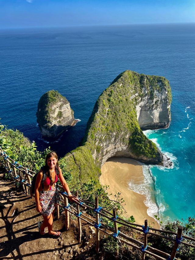 Eine Frau steht auf einem Klippenweg und blickt auf eine einzigartige, grün bewachsene Felsformation und einen Sandstrand mit türkisfarbenem Wasser.