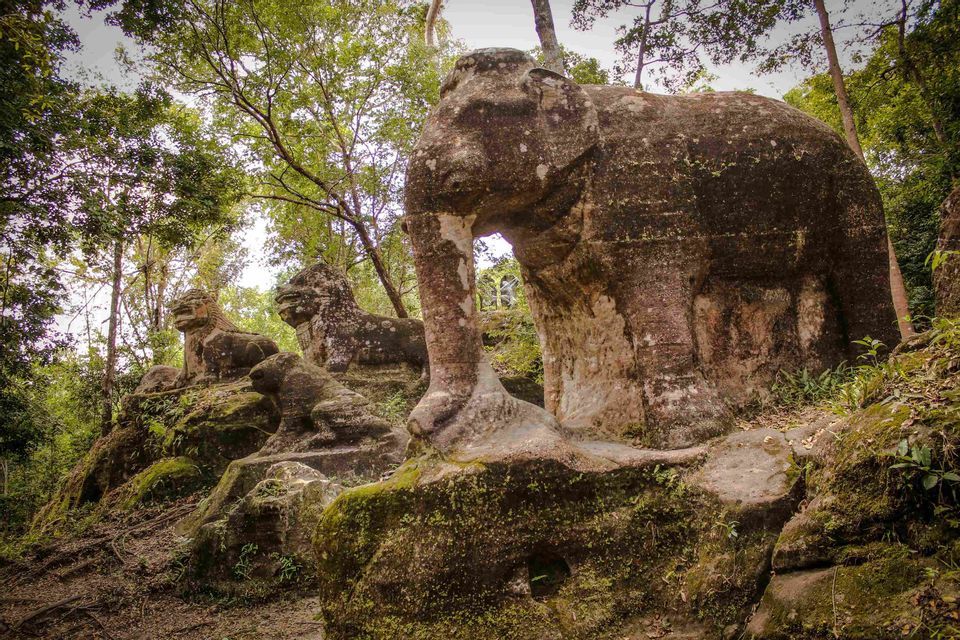 D'anciennes sculptures en pierre recouvertes de mousse représentant un éléphant et des lions se dressent sur un promontoire rocheux dans une jungle verdoyante et dense.
