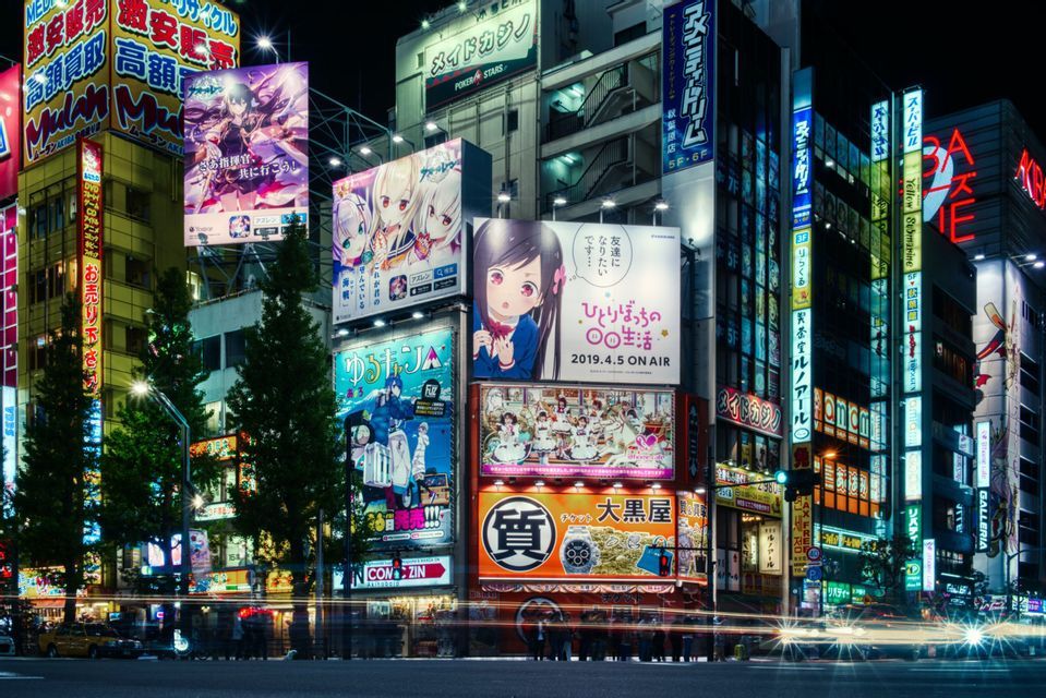 Un'ampia strada cittadina di notte, fiancheggiata da alti edifici coperti da vivaci cartelloni luminosi e insegne al neon con personaggi anime.