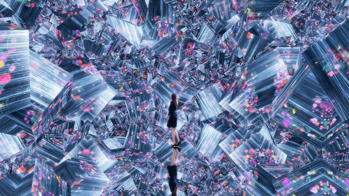 Una donna in abito scuro si trova su una superficie riflettente all'interno di un'installazione caleidoscopica di specchi e luci floreali colorate.