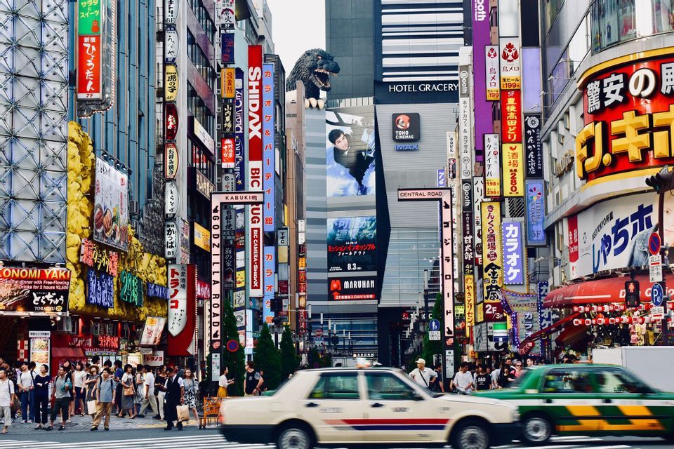 Una strada trafficata in una città giapponese con persone che camminano sui marciapiedi e auto che passano davanti a edifici alti coperti di insegne colorate e una grande testa di Godzilla.