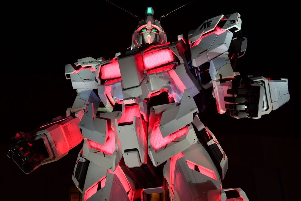 Una statua gigante di robot Gundam bianca illuminata con luci rosse interne contro un cielo notturno scuro.