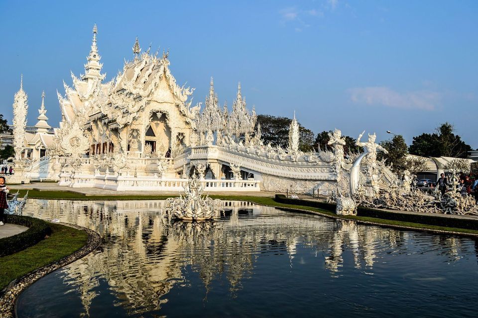 An ornate, all-white temple with detailed spires reflecting in a calm pond under a clear blue sky.