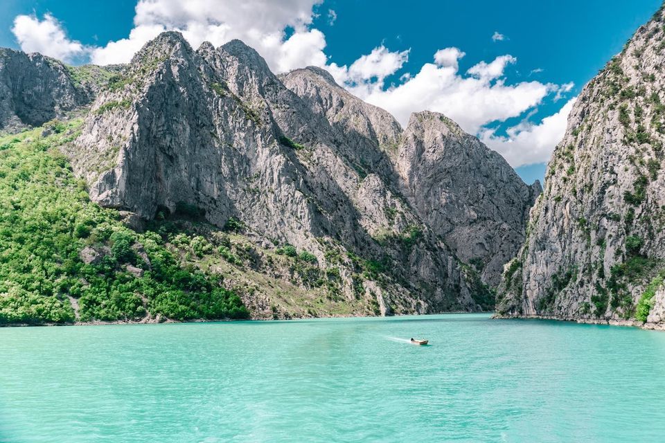 A small boat moves across turquoise water in a canyon surrounded by steep, rocky mountains with green slopes under a blue sky with white clouds.