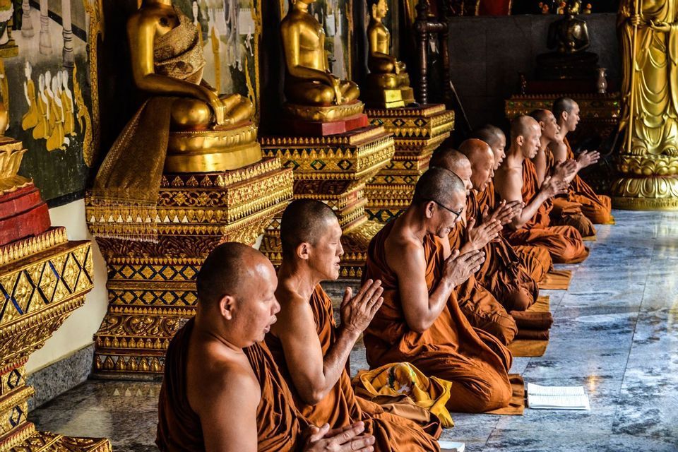 A row of Buddhist monks in orange robes kneeling in prayer inside a temple, with golden Buddha statues in the background.