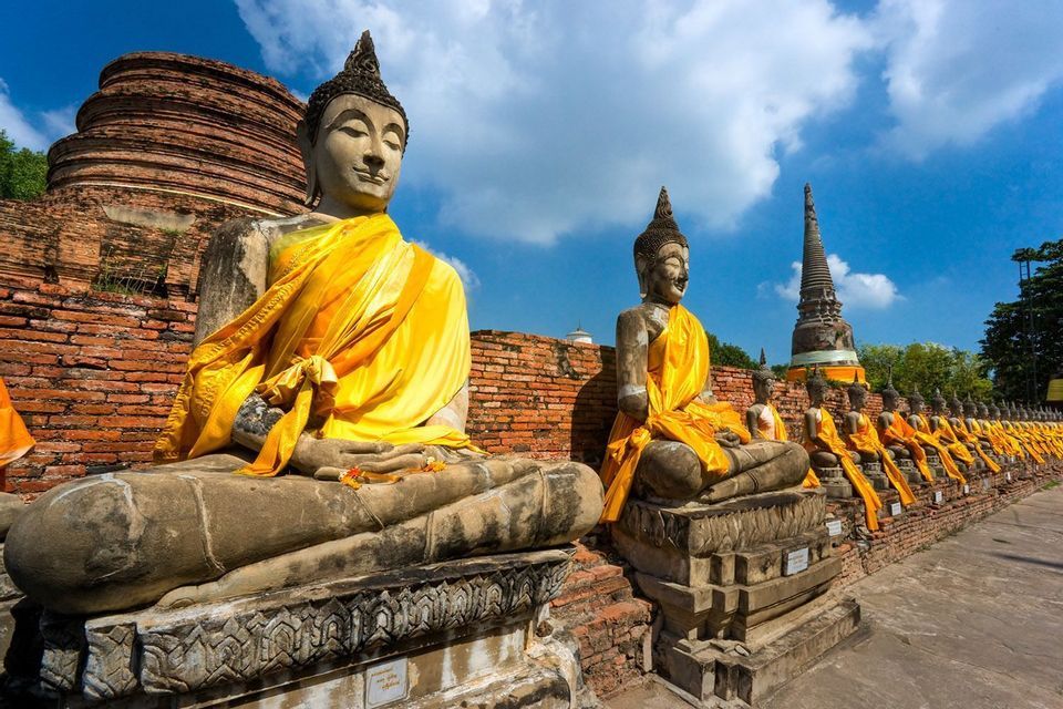 Une rangée d'anciennes statues de Bouddha en pierre, drapées de robes jaune safran, se trouve devant des stupas en brique sous un ciel bleu.