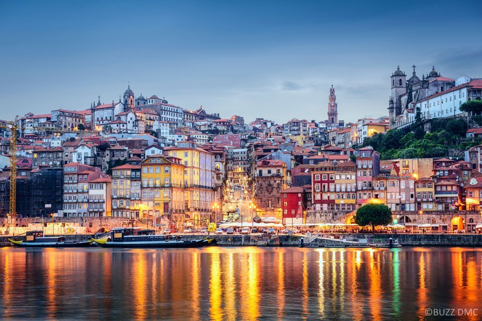 Un denso panorama urbano di edifici colorati su una collina, illuminato al crepuscolo, con riflessi sul fiume in primo piano.