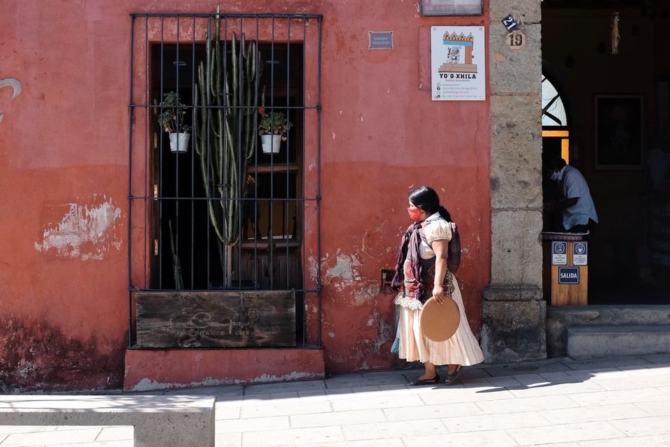 Una mujer con traje tradicional y mascarilla camina por una acera junto a un edificio rojo-naranja con una ventana enrejada.