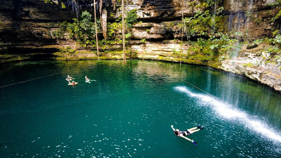 Vista aérea de un viaje en grupo de WeRoad nadando y flotando en el agua turquesa de un cenote rodeado de acantilados rocosos.