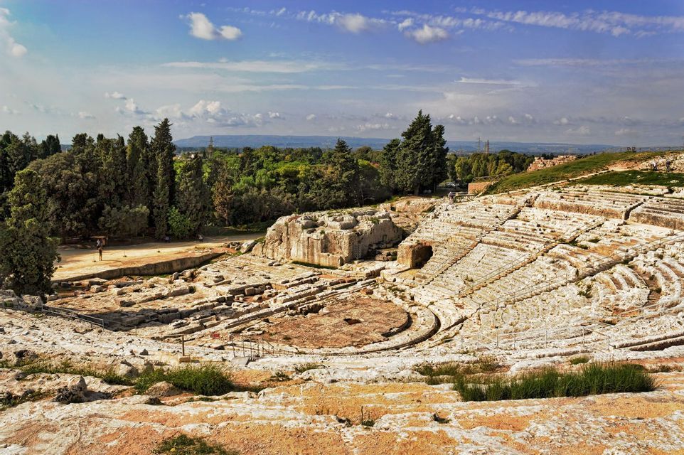 The stone ruins of an ancient amphitheater are built into a hillside, surrounded by green trees under a blue sky.