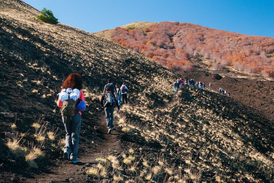 A WeRoad group trip hiking in a single file line up a narrow dirt trail on a steep, rocky mountainside under a clear blue sky.