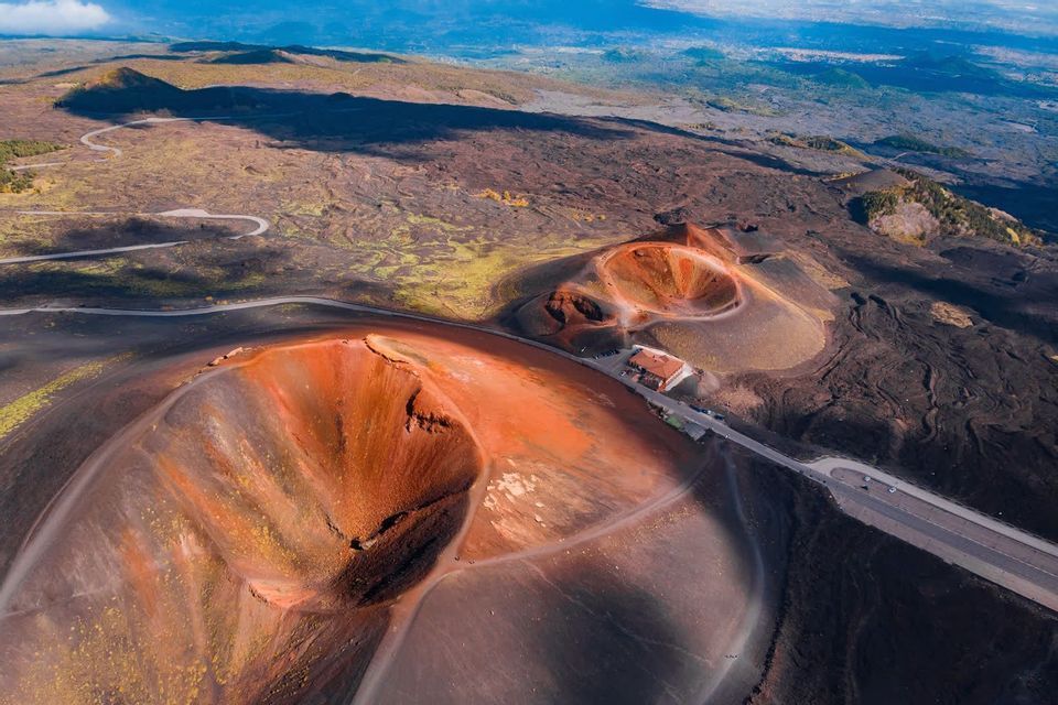 Una vista aerea di crateri vulcanici rossastri e una strada tortuosa che attraversa un paesaggio roccioso e scuro sotto un cielo azzurro.