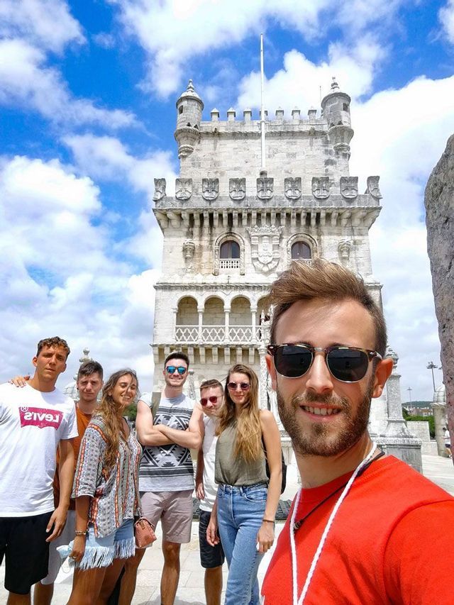 Un uomo con occhiali da sole si scatta un selfie con un gruppo WeRoad di fronte a un'antica torre di pietra sotto un cielo azzurro e nuvoloso.