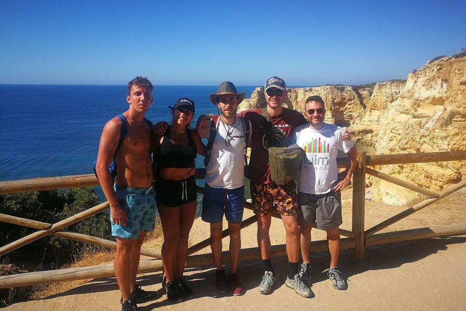 Un voyage de groupe WeRoad de cinq personnes posant pour une photo sur un point de vue côtier ensoleillé, avec l'océan bleu et des falaises en arrière-plan.