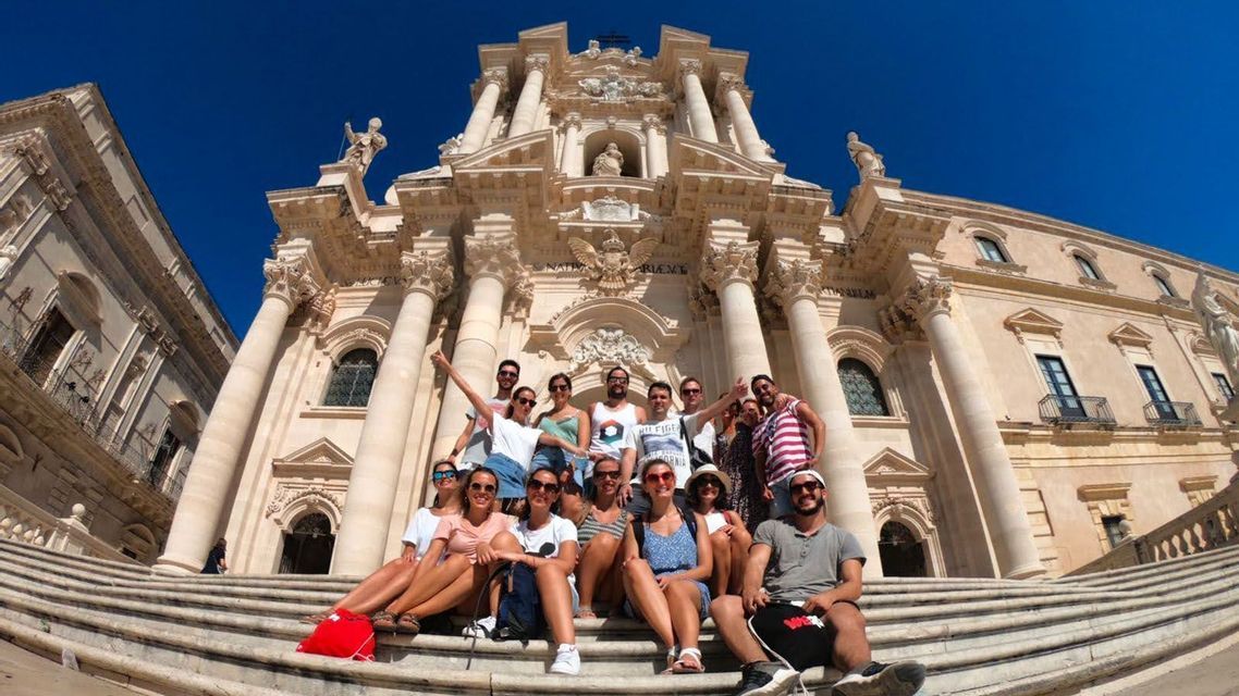 Un grupo de viaje de WeRoad posando para una foto en las escalinatas de un edificio barroco ornamentado bajo un cielo azul despejado.