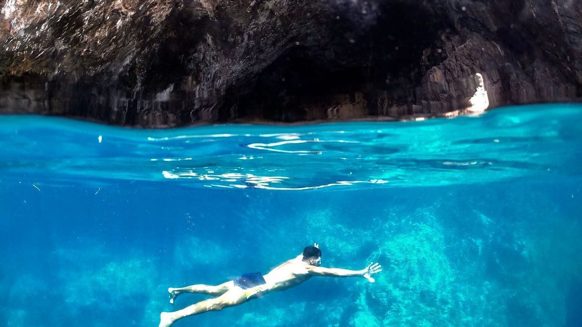Un uomo nuota sott'acqua in acque cristalline e blu, con la scura parete rocciosa di una grotta marina visibile sopra la superficie.