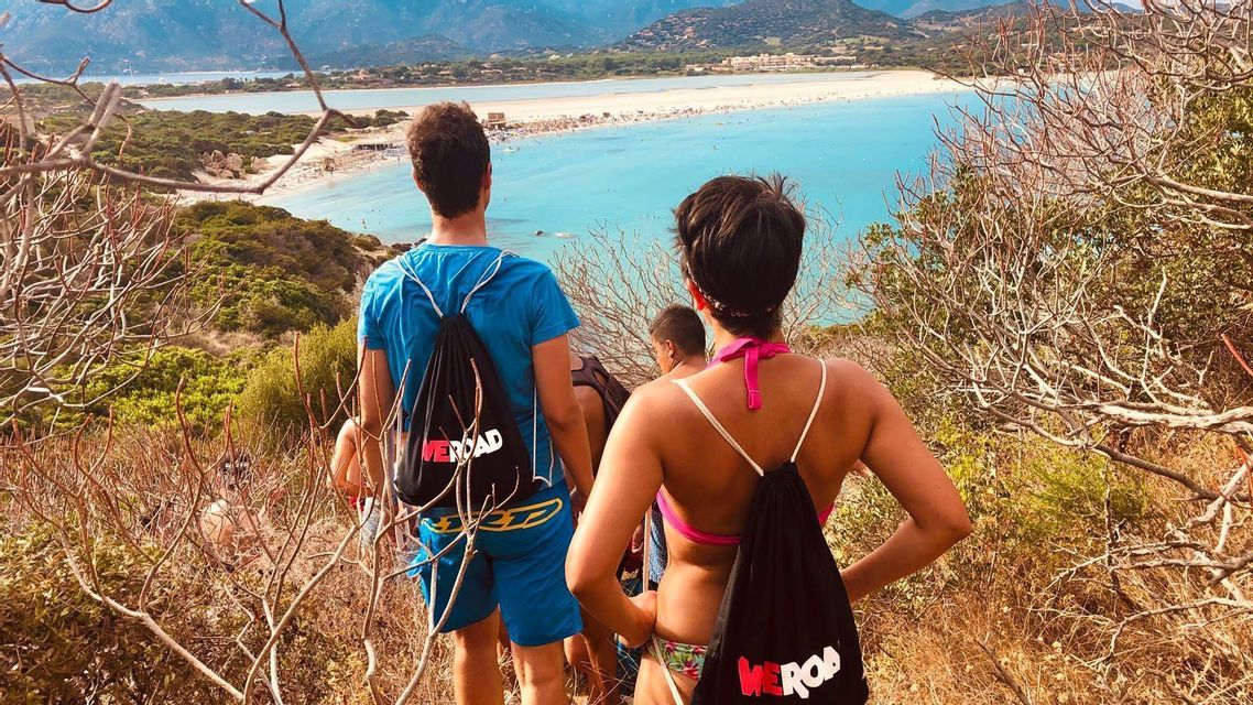 Un gruppo WeRoad, visto da dietro, si erge su una collina che si affaccia su una spiaggia affollata con acqua turchese brillante.