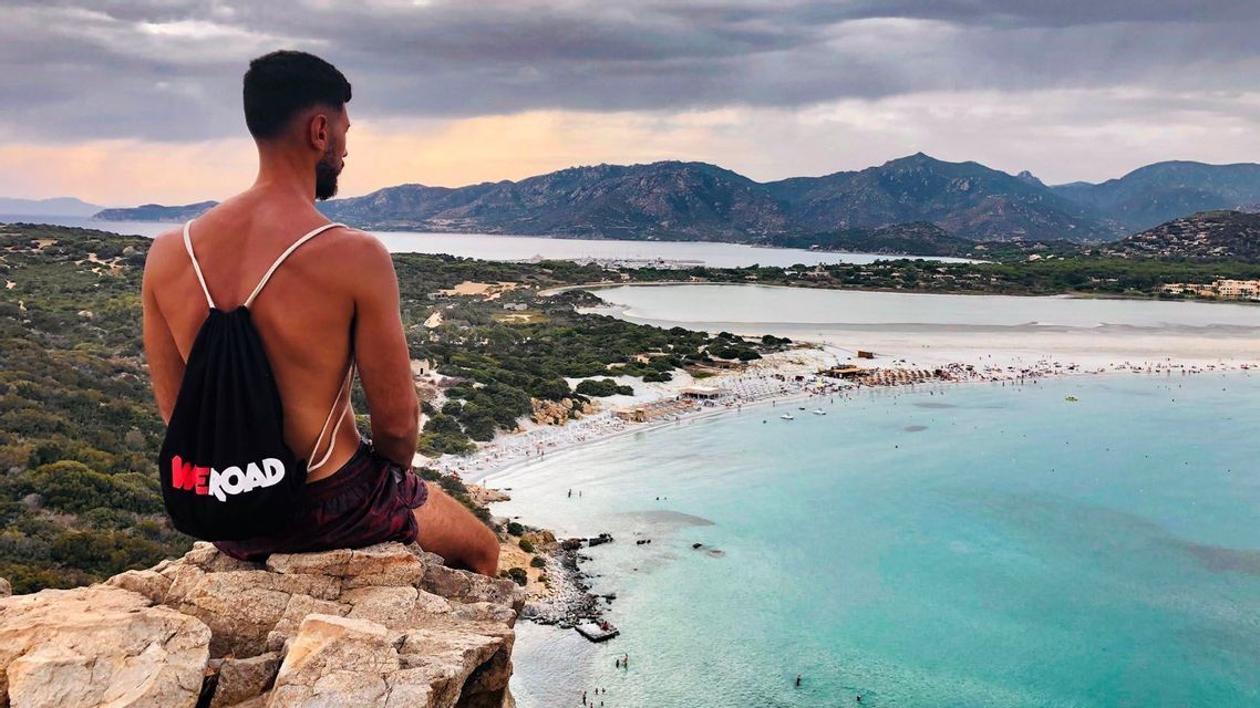 Un uomo con uno zaino WeRoad siede su una scogliera rocciosa, ammirando una spiaggia affollata con acqua turchese e montagne.