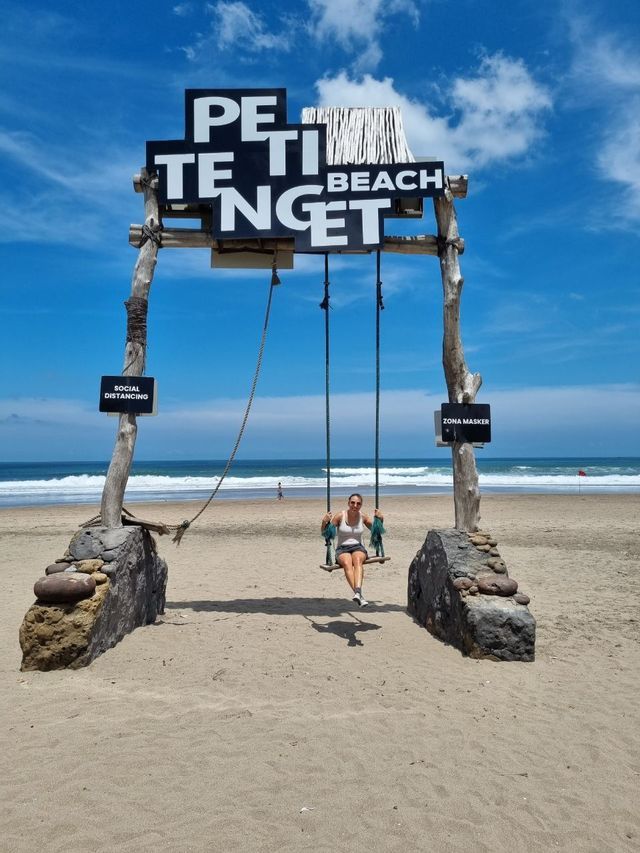 Eine Frau sitzt auf einer großen Holzschaukel an einem Sandstrand unter einem Schild mit der Aufschrift 'Petitenget Beach', mit dem Meer im Hintergrund.