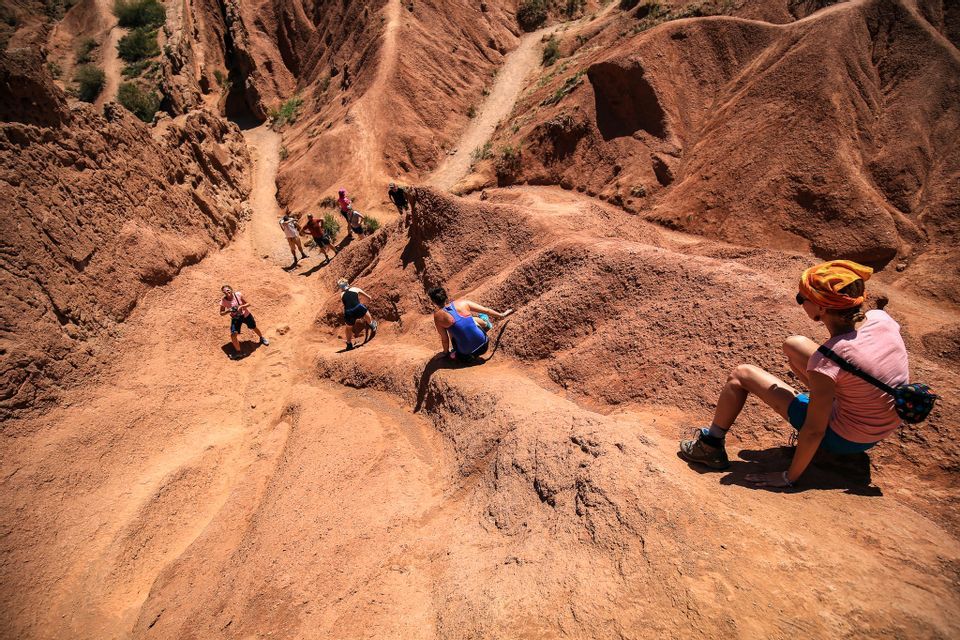 Un viaggio di gruppo WeRoad percorre uno stretto sentiero attraverso un ripido canyon di roccia rossa sotto il sole splendente.