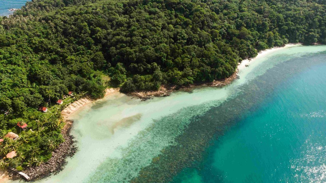 Vista aerea di un'isola tropicale lussureggiante con piccole case adagiate su una spiaggia sabbiosa e acqua turchese cristallina.