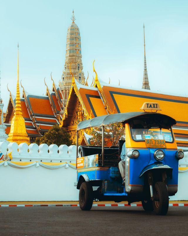 Un tuk-tuk blu e giallo parcheggiato su una strada davanti a un tempio decorato con guglie dorate e tetti arancioni.