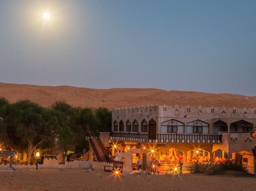 Un lodge illuminato nel deserto con alberi si annida tra le dune di sabbia sotto una luna piena al crepuscolo.