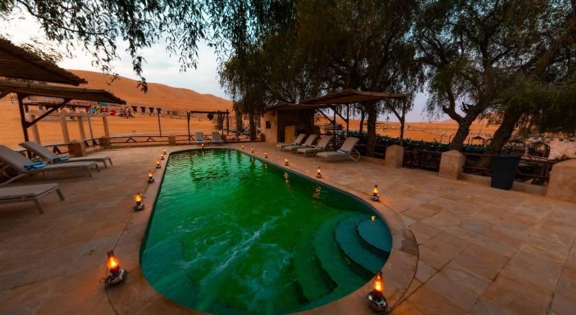Una piscina con acqua verde, illuminata da piccole lanterne, si trova in un'oasi nel deserto con sedie a sdraio e dune di sabbia sullo sfondo.