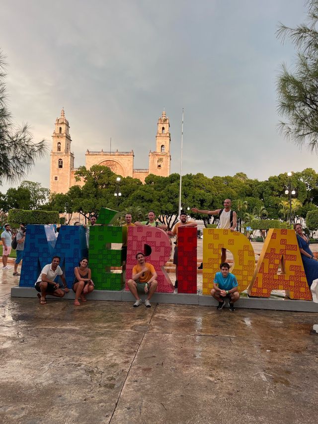 Un viaggio di gruppo WeRoad in posa per una foto con le grandi e colorate lettere che formano la parola MERIDA in una piazza con una cattedrale sullo sfondo.