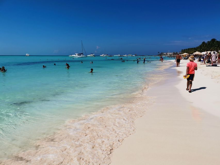 Muchas personas disfrutan de un día soleado en una playa con arena blanca y agua turquesa cristalina, con varios barcos en el horizonte.