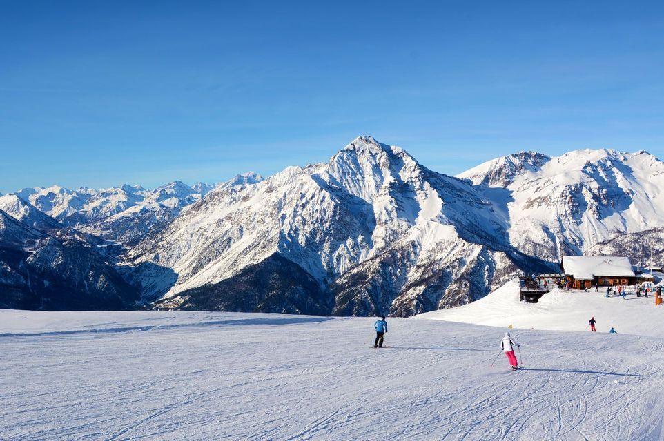 Personen beim Skifahren auf einer breiten, verschneiten Piste mit einer riesigen Bergkette und einer Skihütte im Hintergrund unter klarem blauem Himmel.