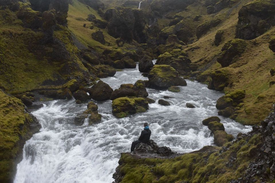 Una persona se sienta en una roca cubierta de musgo con vistas a los rápidos de un río caudaloso en un cañón verde y escarpado.