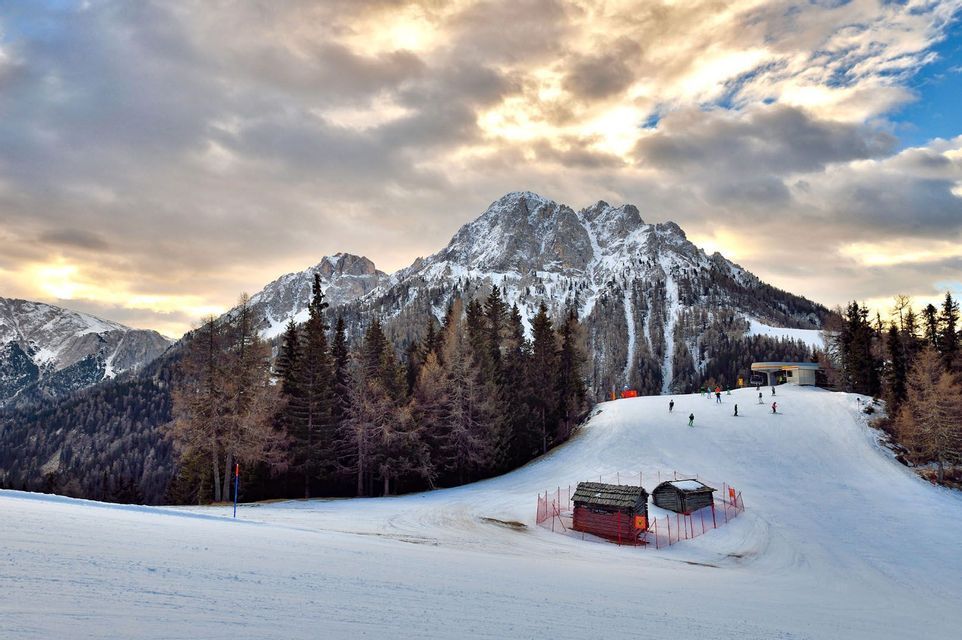 Ein schneebedeckter Berggipfel ragt über einer Skipiste auf, wo Menschen an einem Wald und kleinen Holzhütten unter einem bewölkten Himmel vorbeifahren.