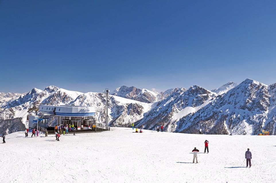 Personen beim Skifahren auf einer breiten, verschneiten Piste neben einer Skiliftstation, mit einer Reihe schneebedeckter Berge im Hintergrund.
