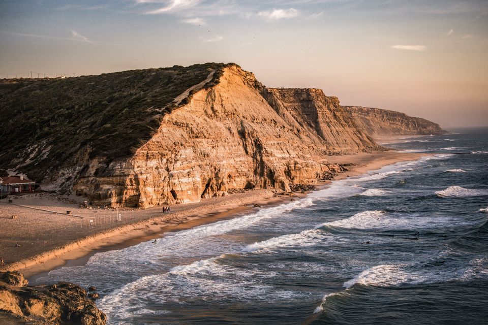 Eine Luftaufnahme einer Küste mit steilen, felsigen Klippen, die von der Sonne beleuchtet werden, einem Sandstrand darunter und dunklen Meereswellen mit weißem Schaum.