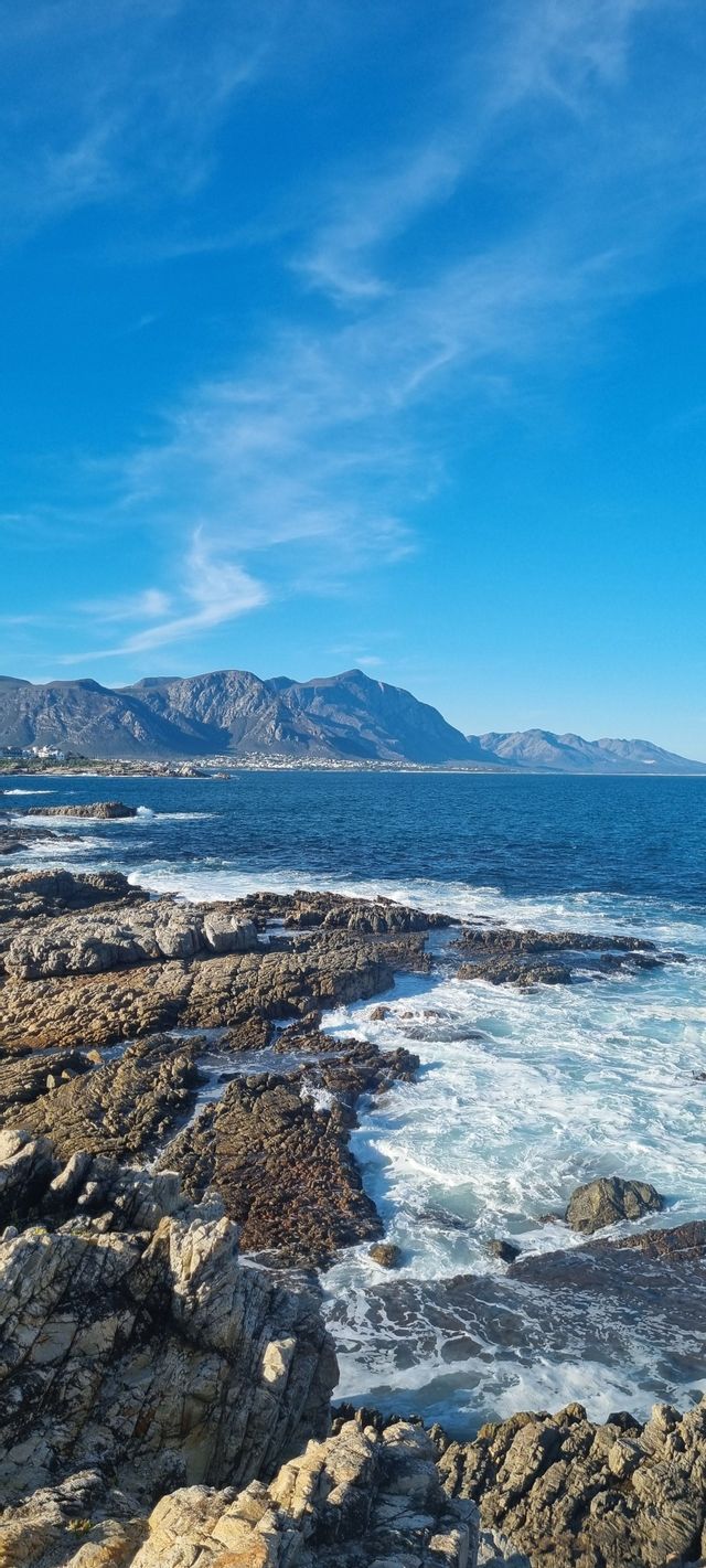 Le onde dell'oceano si infrangono contro una costa rocciosa con montagne aspre che si estendono lungo l'orizzonte sotto un cielo azzurro e limpido.