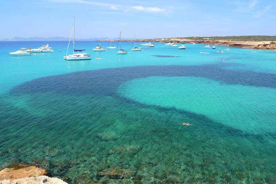 An aerial view of a calm bay with numerous boats anchored in clear, turquoise water over a rocky seabed.