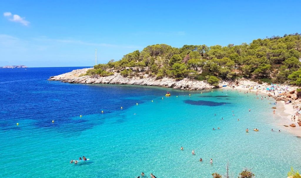 People swimming and relaxing in a cove with clear turquoise water and a sandy beach, backed by a tree-covered rocky hill.