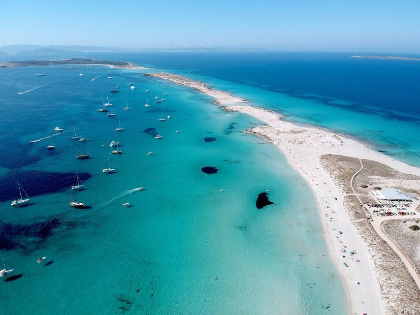 An aerial view of numerous sailboats anchored in clear turquoise waters alongside a long white sand spit.