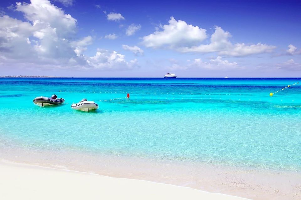Two dinghies float on calm turquoise water near a white sand beach, with a cruise ship on the horizon.
