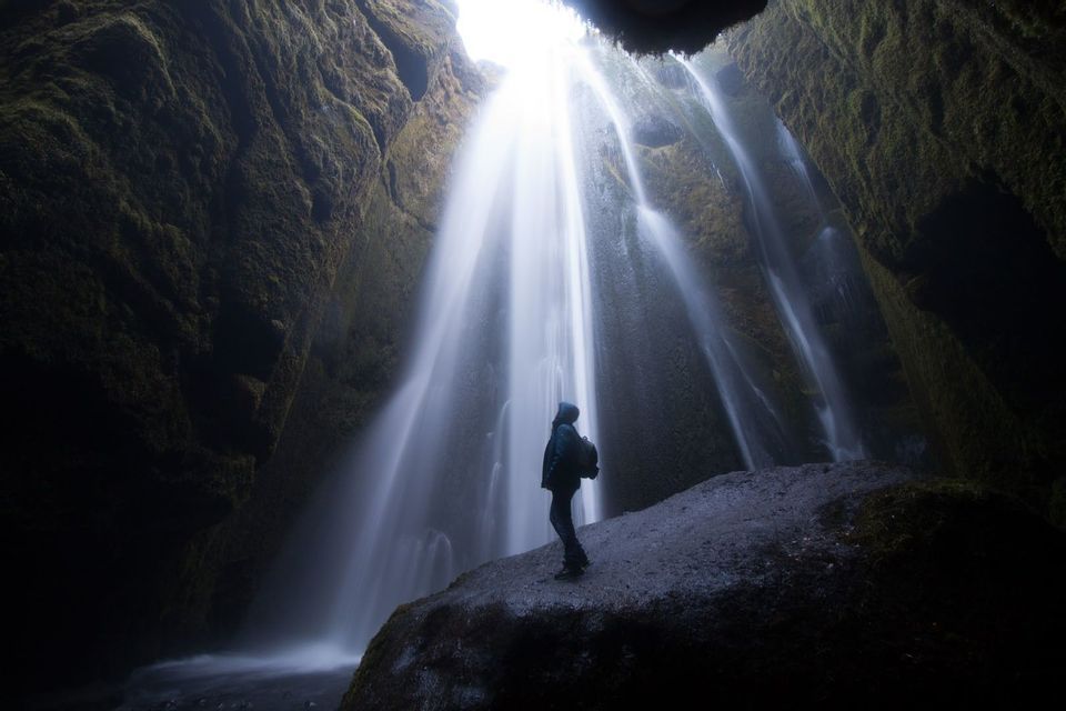 Una persona silueteada con una mochila de pie sobre una roca dentro de una cueva musgosa, mirando una potente cascada.